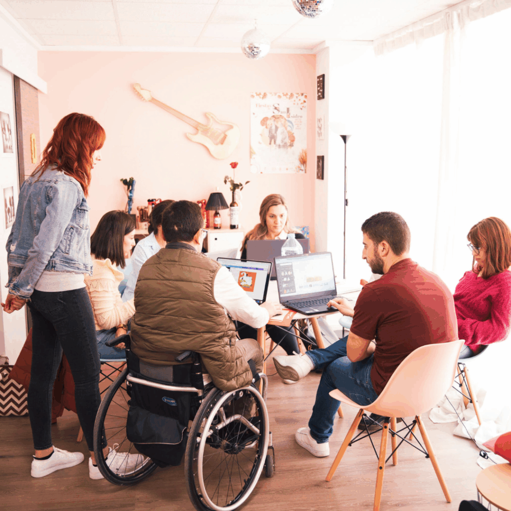 Diverse group of professionals working together around laptops, including a person in a wheelchair, collaborating in a modern office setting.